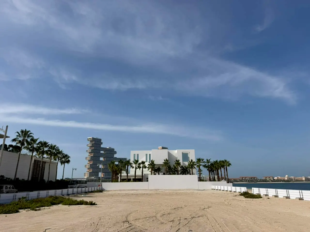 Beachfront property with modern white buildings, palm trees, and blue sky. Sandy lot in foreground.