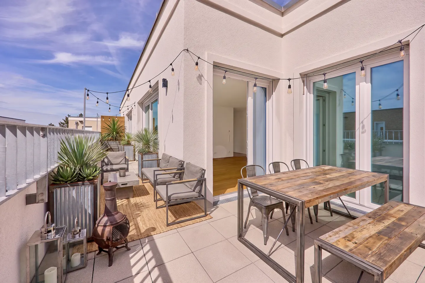 Modern rooftop terrace with a wooden dining set, lounge seating, string lights, and potted plants under a blue sky.