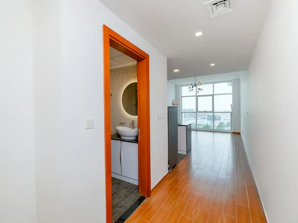 Interior view of a modern apartment with white walls, wood floors, and an orange-framed bathroom doorway. A sink and round mirror are visible in the bathroom.