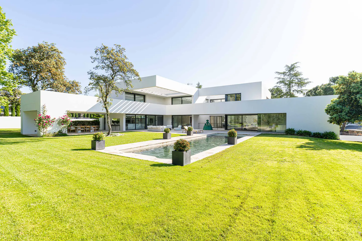 Modern white house with a rectangular pool in the backyard, surrounded by green grass and trees under a clear blue sky.