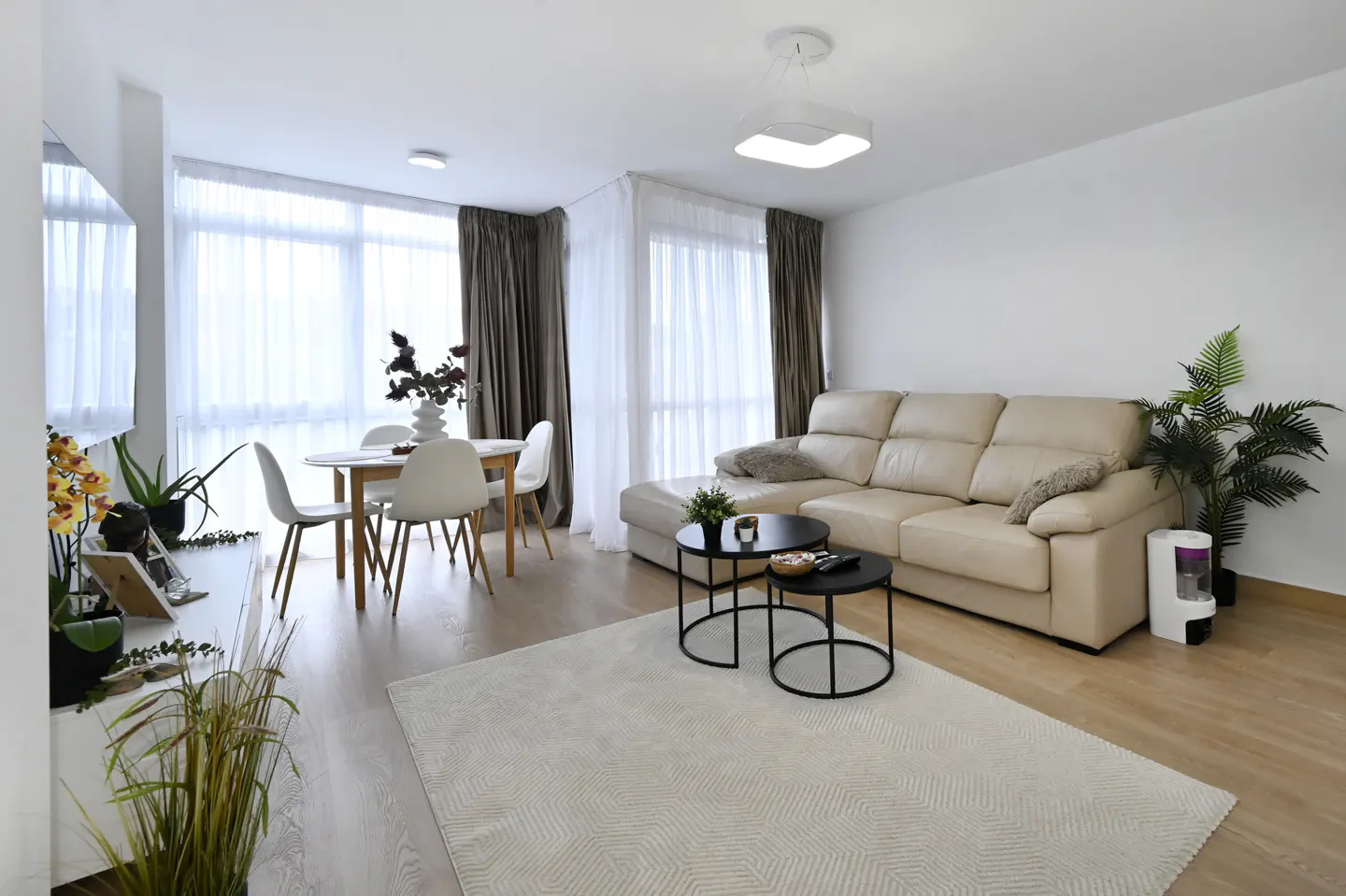 Bright living room with a beige sectional sofa, black nesting tables, and a patterned rug. A dining table with white chairs sits near sheer-curtained windows.