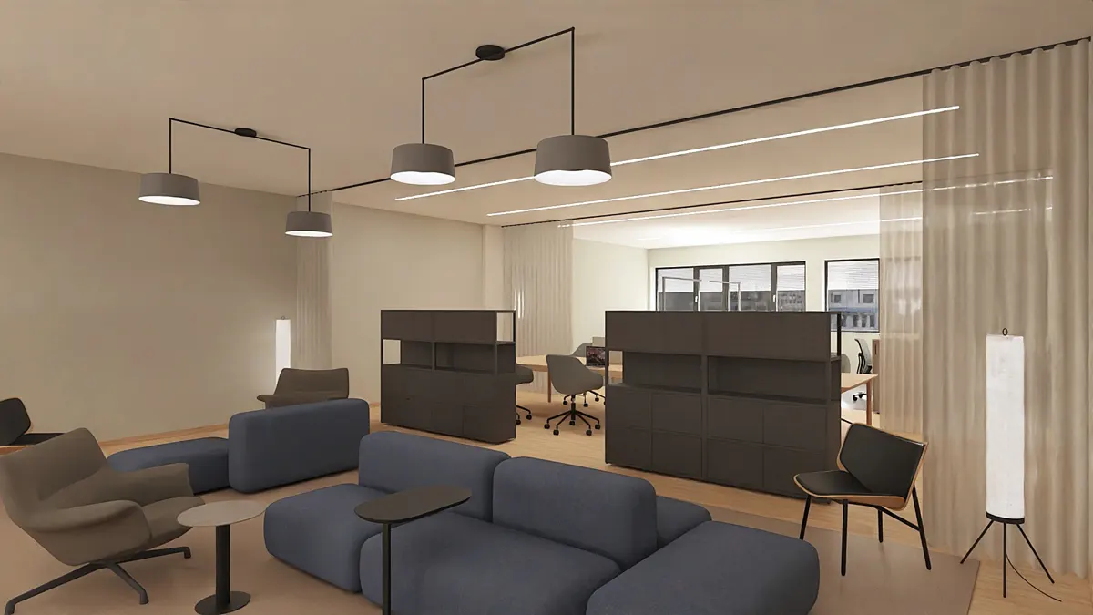 Modern office space with blue sofas, grey chairs, and dark grey shelving units. Pendant lights hang from the ceiling. A large window is visible in the background.