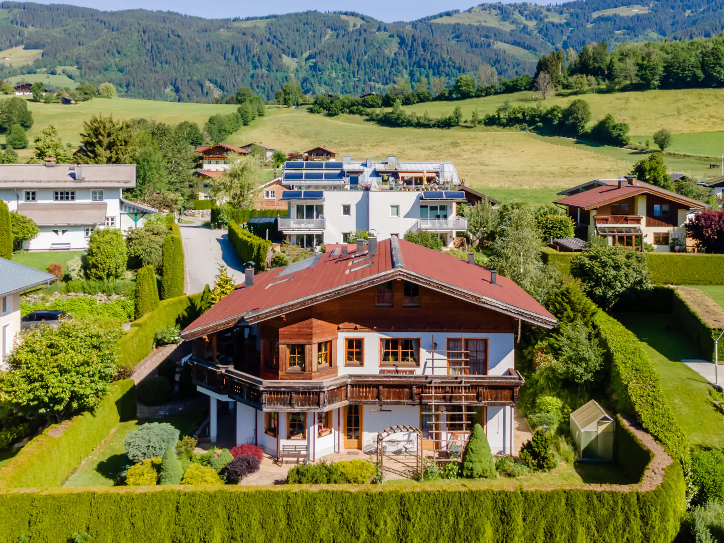 Aerial view of a traditional wooden chalet with a red roof, surrounded by lush green hedges and scenic mountain landscapes.
