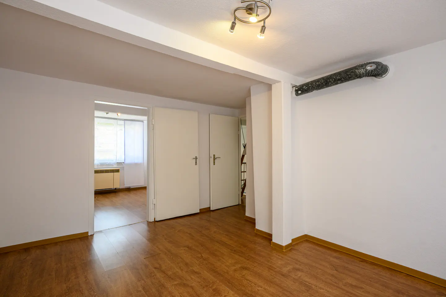 A bright room with wood floors, white walls, and a ceiling light. Two white doors are visible, one leading to another room with a window.