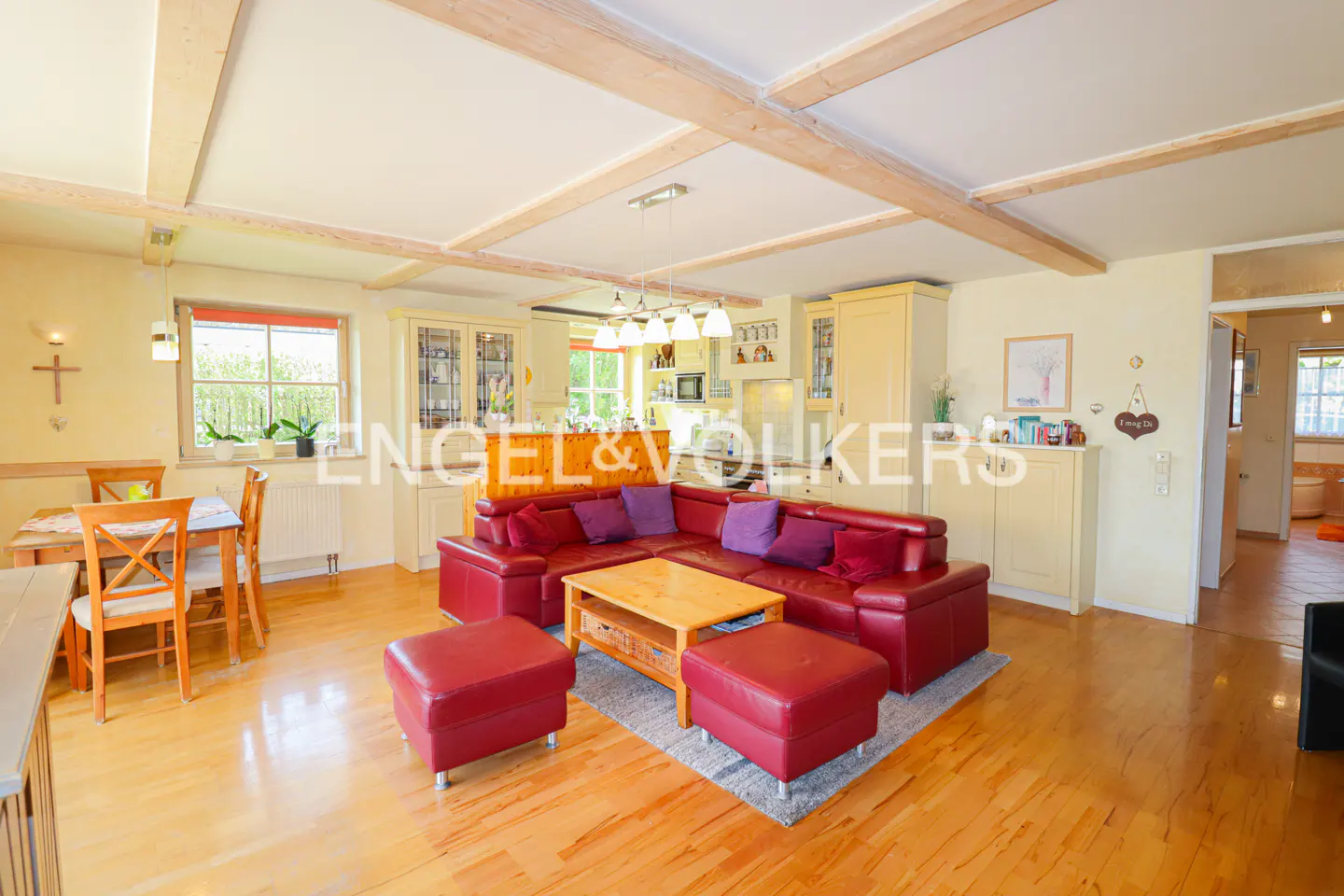 Open-concept living space with a red leather sectional, wood floors, and exposed beams. Kitchen and dining area visible.
