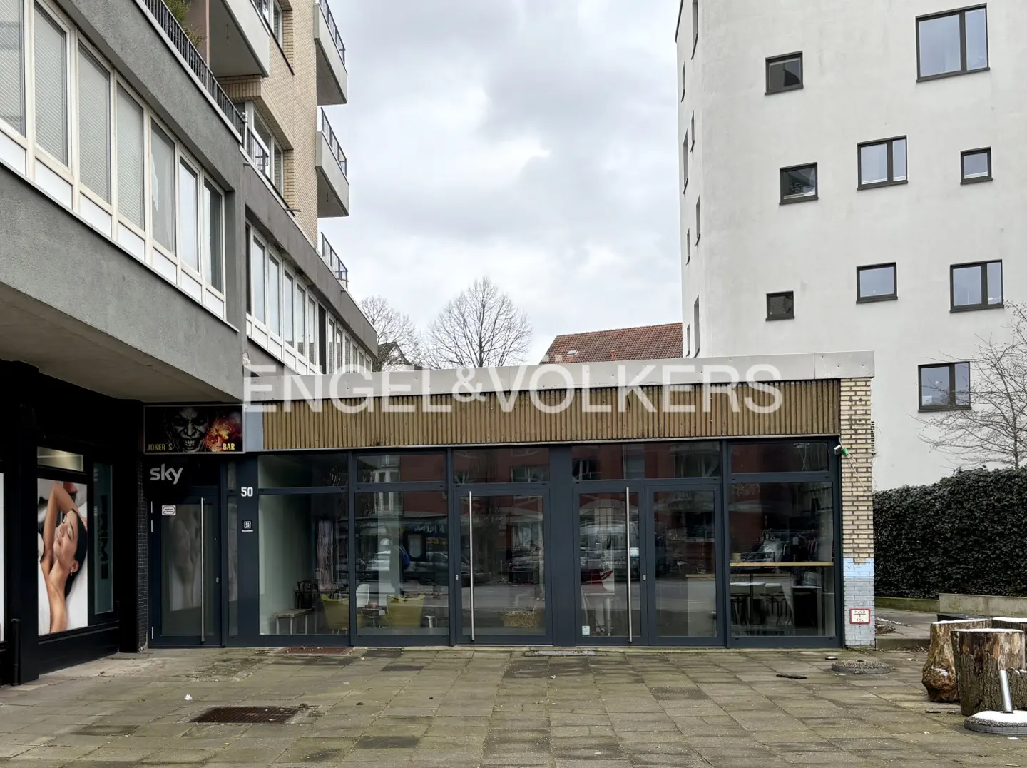 Engel & Völkers storefront with large windows and gray frames, between two buildings on a cloudy day.