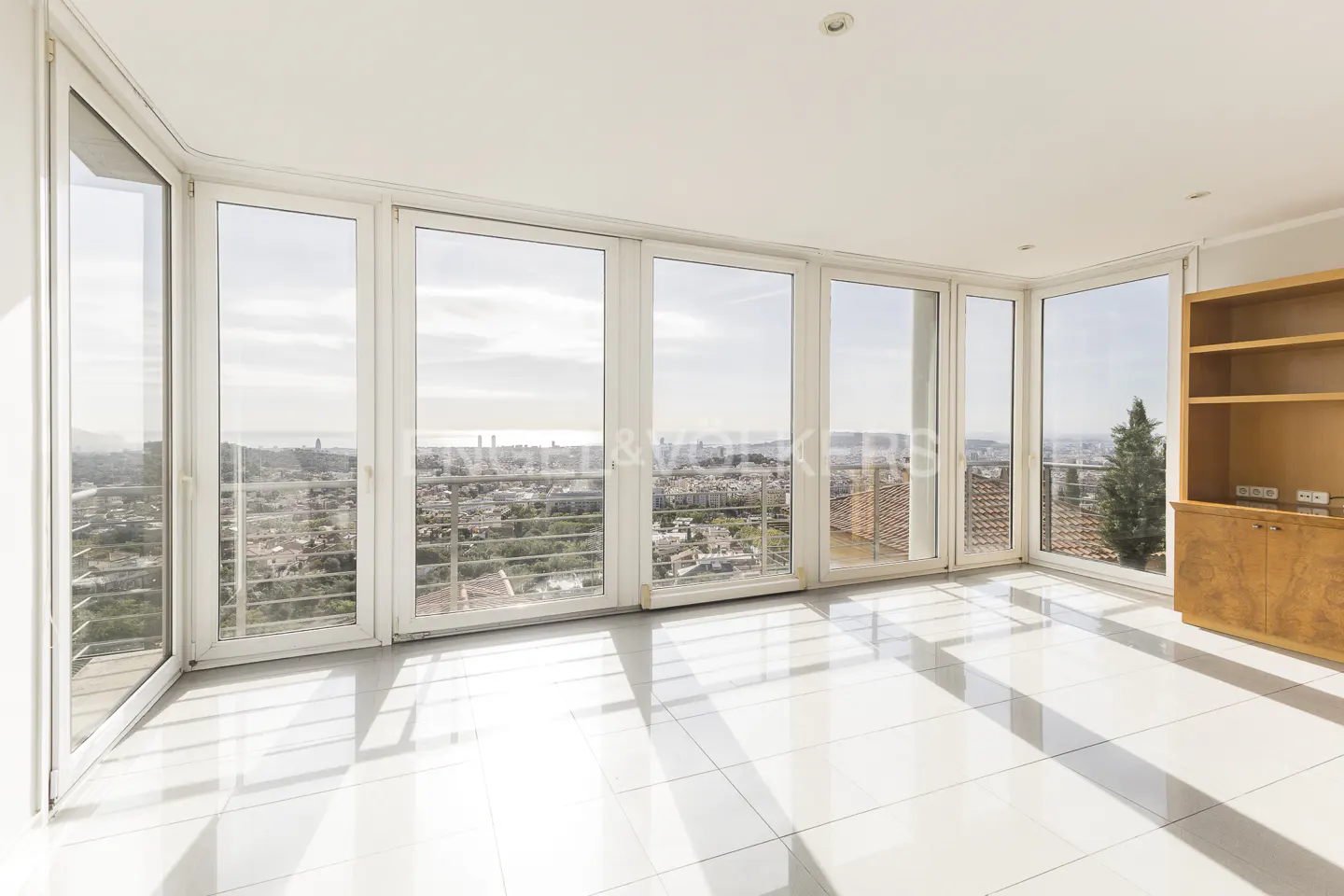 Bright, empty room with white tile floor and large windows showing a city view. A wooden cabinet stands against the right wall.