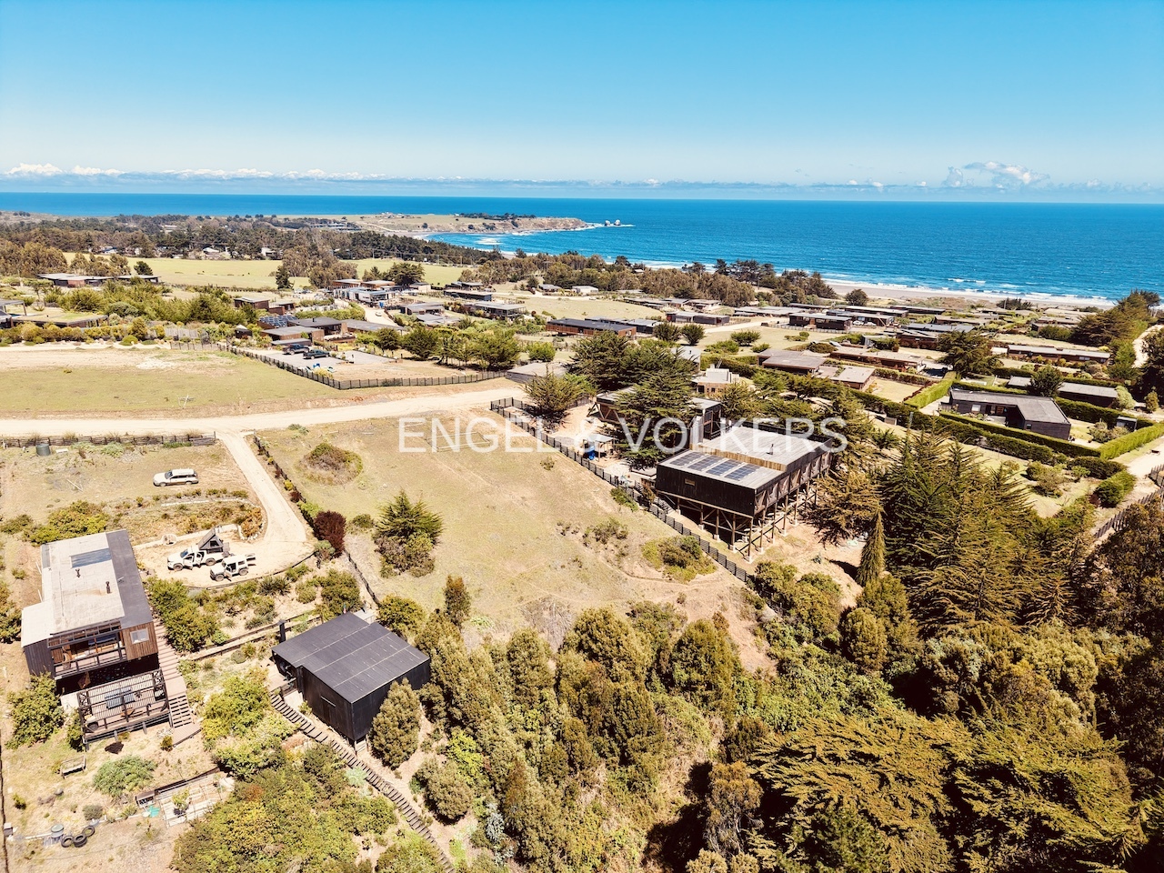 Aerial view of modern black houses nestled in a coastal landscape with trees, rolling hills, and the blue ocean beyond. [AI-generated text]