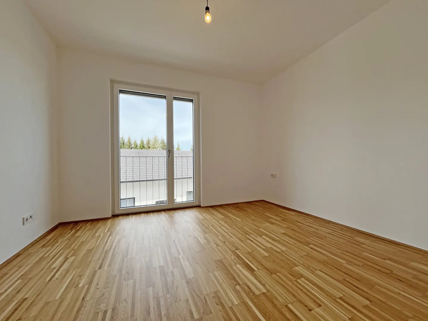Empty room with light wood floors, white walls, and a sliding glass door to a balcony. A single lightbulb hangs from the ceiling.