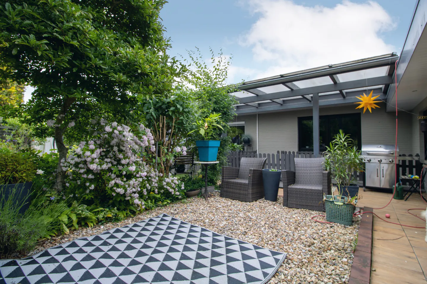 A gravel patio with a black and white geometric rug, two wicker chairs, a grill, and lush garden plants under a pergola.