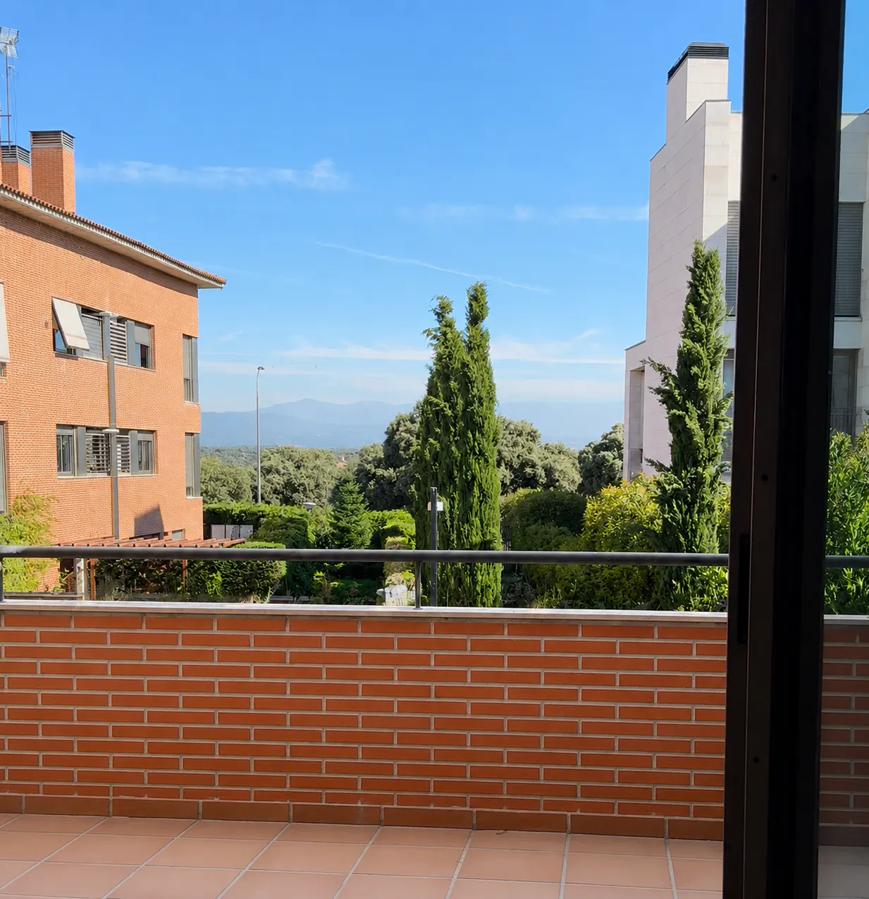 View from a balcony with a brick railing overlooking a garden, trees, and neighboring apartment buildings under a blue sky.