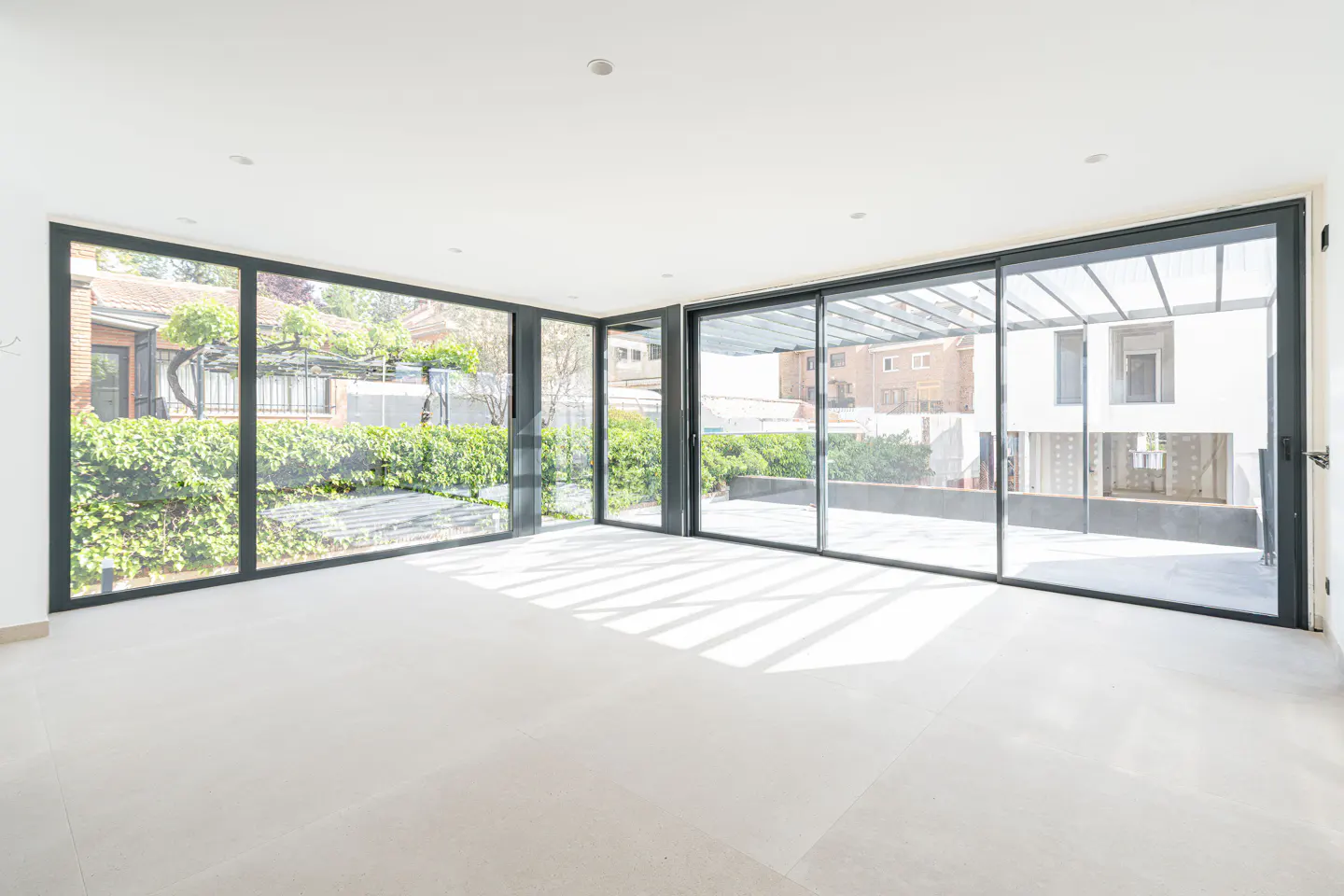 Bright, empty room with white walls and floor. Large black-framed windows offer views of green foliage and nearby buildings. Sunlight streams in.