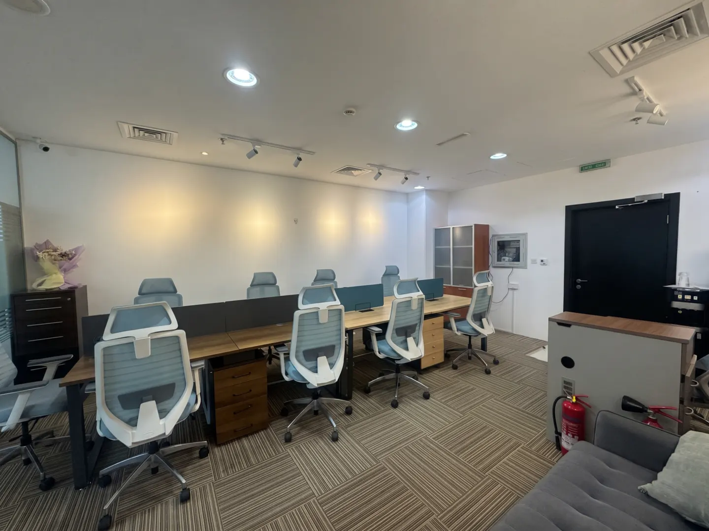 Bright office space with desks, blue chairs, and a striped carpet. A black door and gray sofa are visible.