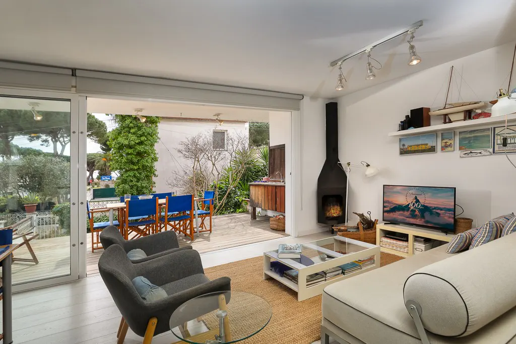 Modern living room with a fireplace, sofa, and armchairs, opening onto a wooden deck with blue dining chairs and greenery.