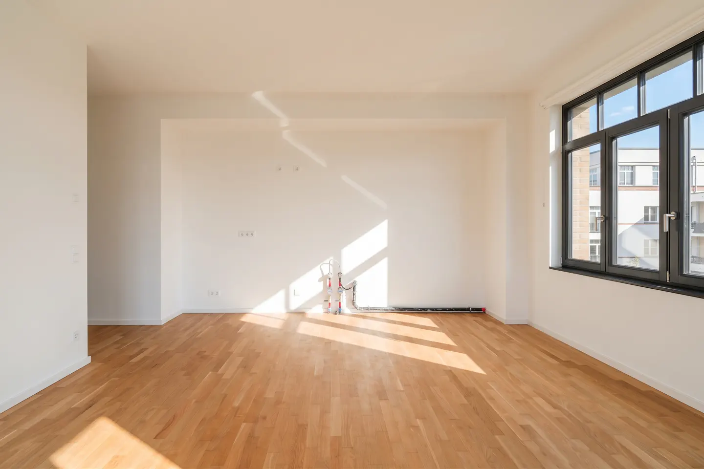 Bright, empty room with light wood floors, white walls, and a large black window. Sunlight streams in, casting shadows across the floor.