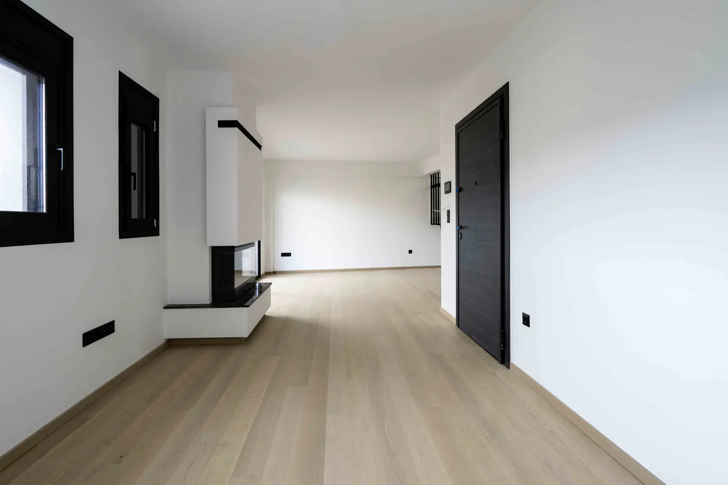Bright, empty room with light wood floors, white walls, and black trim. A modern fireplace is on the left, and a dark door is on the right.