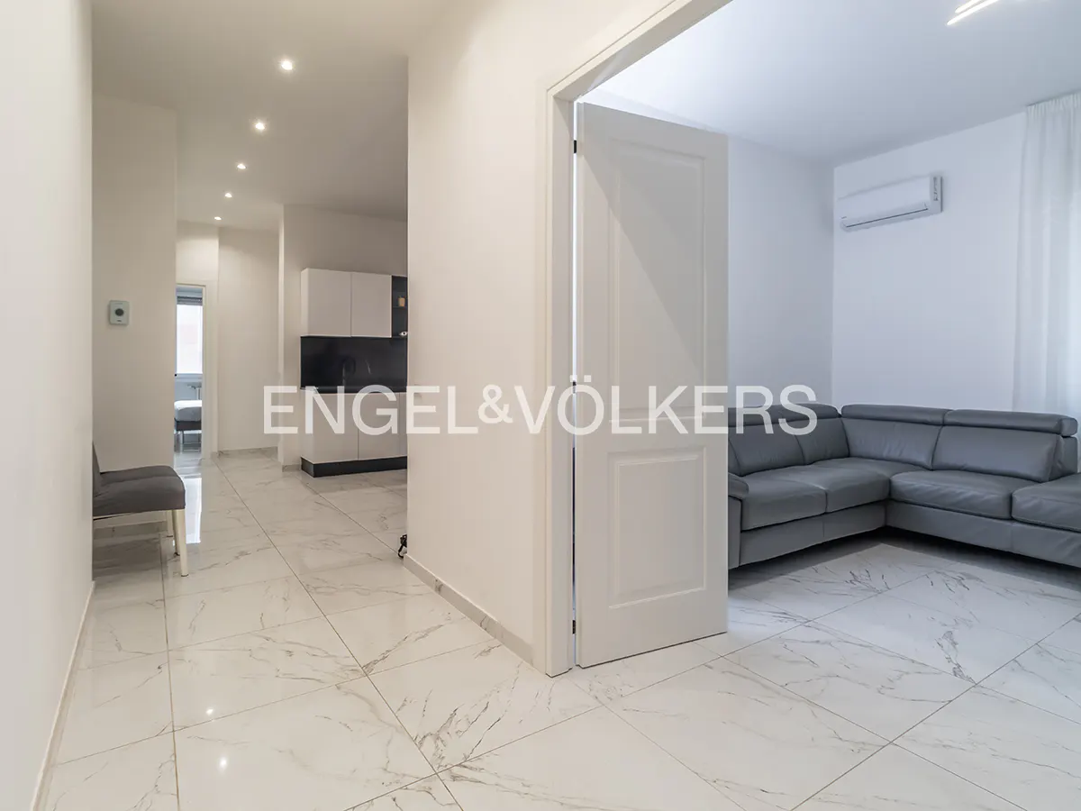 A bright, modern apartment interior with white walls, marble floors, and a gray sectional sofa. A kitchen is visible in the background.