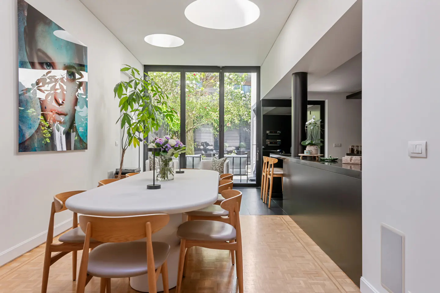 Bright dining room with white walls, parquet floors, and a white oval table surrounded by wooden chairs. A large painting hangs on the wall. Black kitchen in the background.