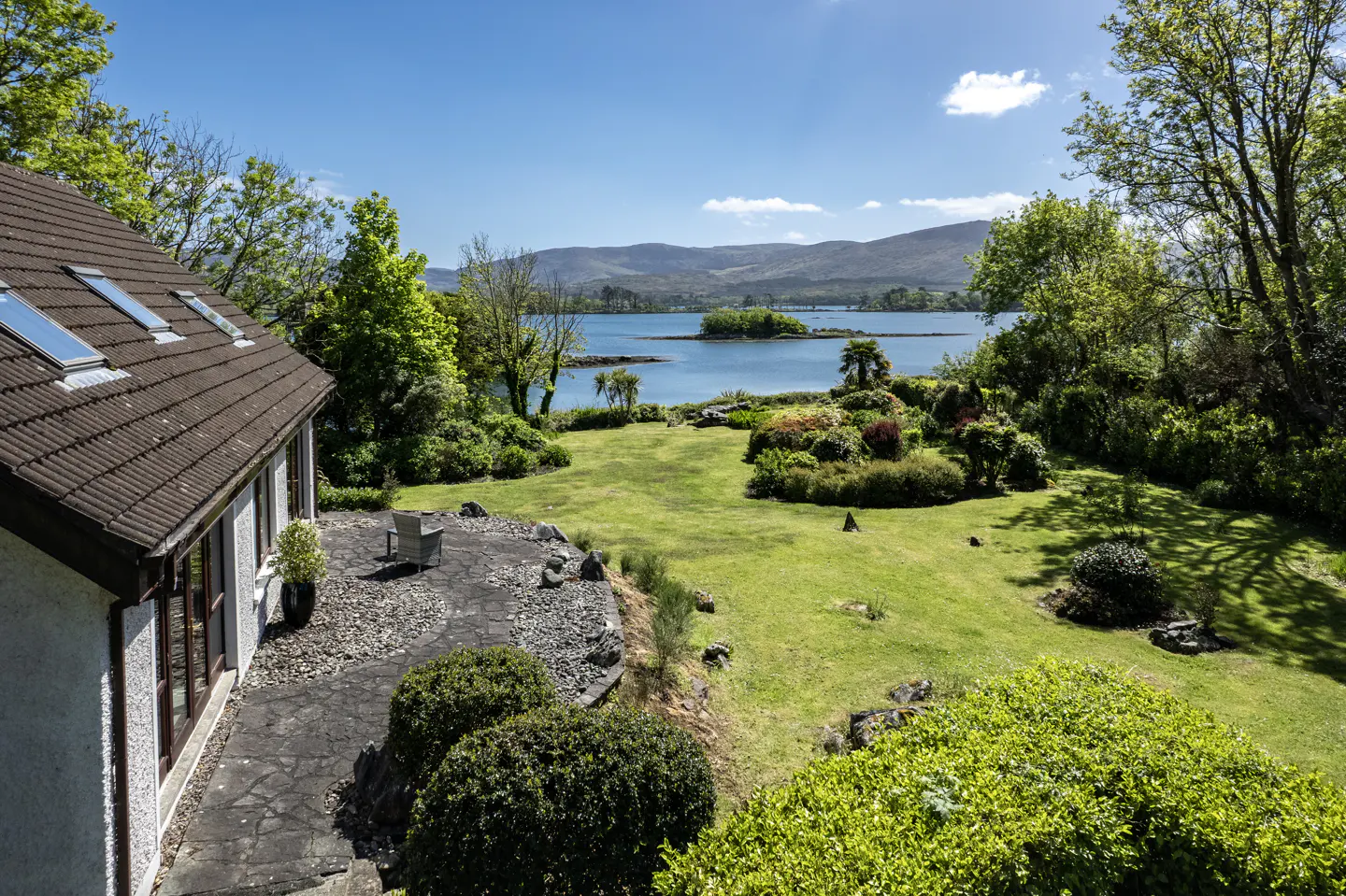 Exterior view of a house with a brown roof, a stone patio, and a green lawn overlooking a lake with islands and mountains in the background.