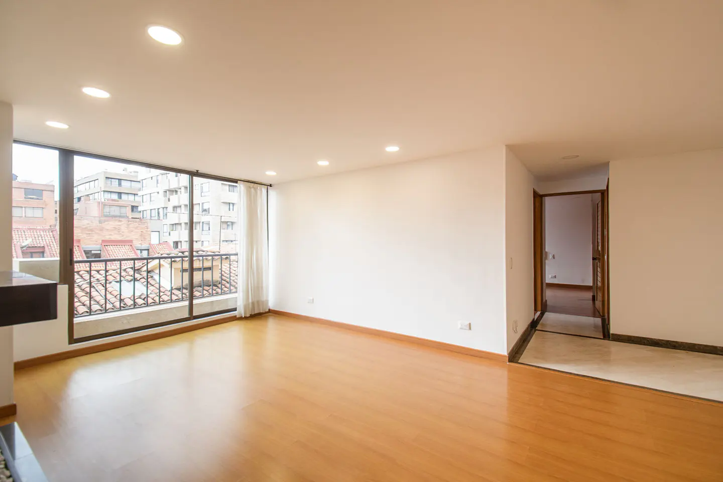 Bright, empty room with wood floors, white walls, and a large window with a city view. An open doorway leads to another room.