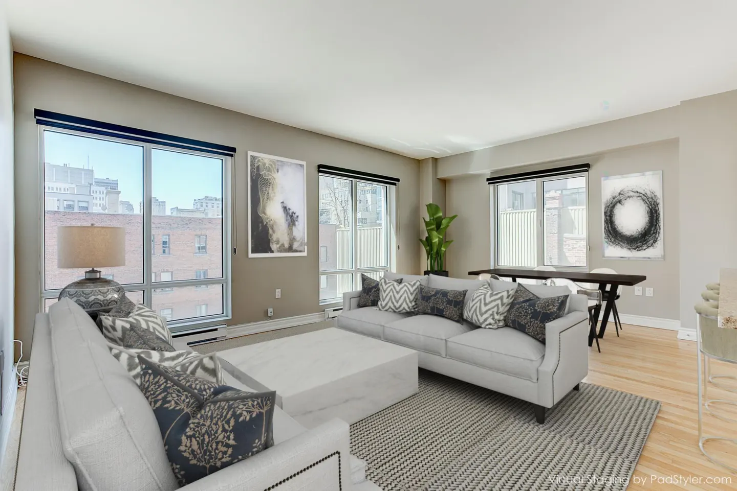 Bright living room with white sofas, patterned pillows, and a large white ottoman on a gray rug. Windows offer city views.