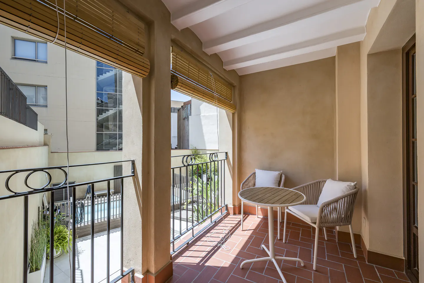 A sunlit balcony with terracotta tiles, a small table, and two chairs. Woven blinds are partially drawn, revealing a view of a building and pool.