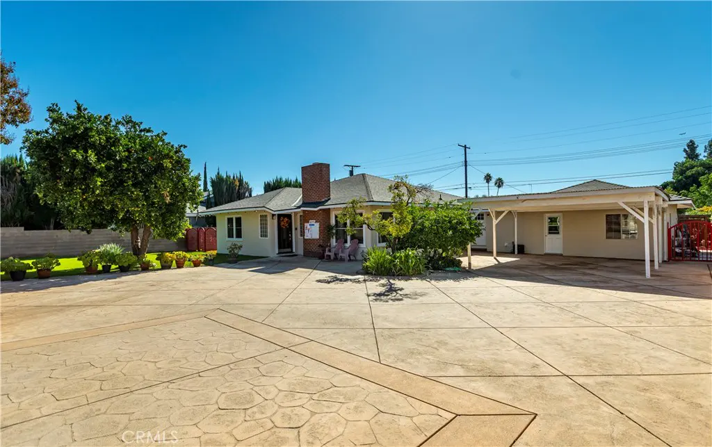 A tan single-story house with a brick chimney, a large driveway, and a carport on a sunny day.