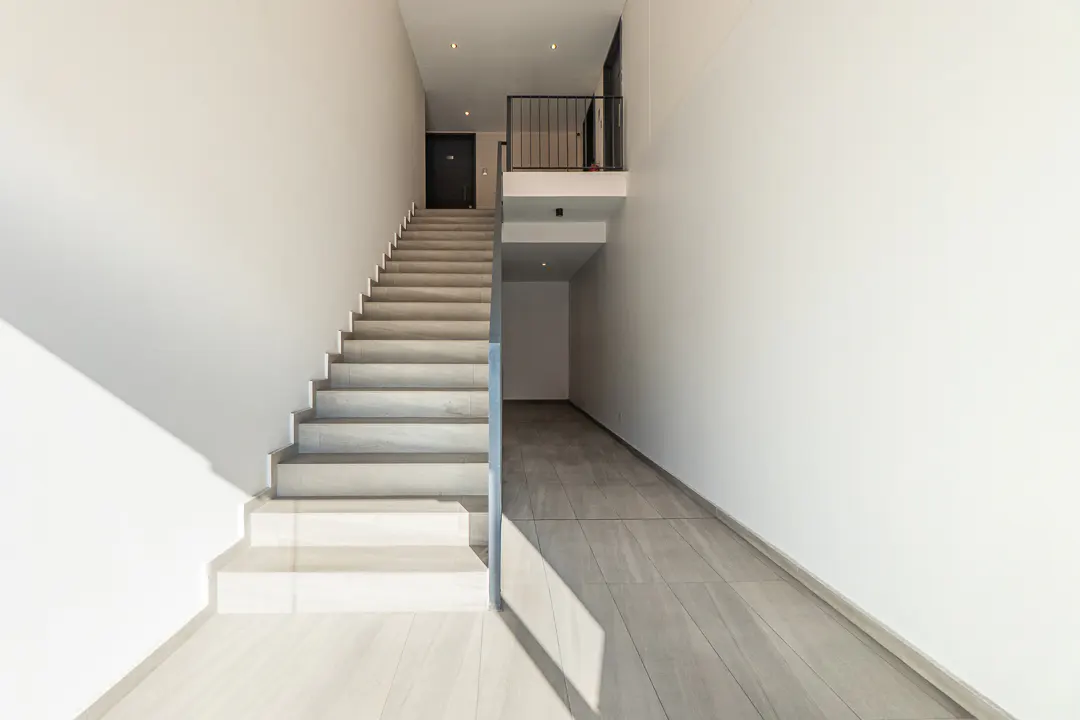 Modern building interior with white walls, gray tile floor, and a staircase leading to a second-floor balcony.