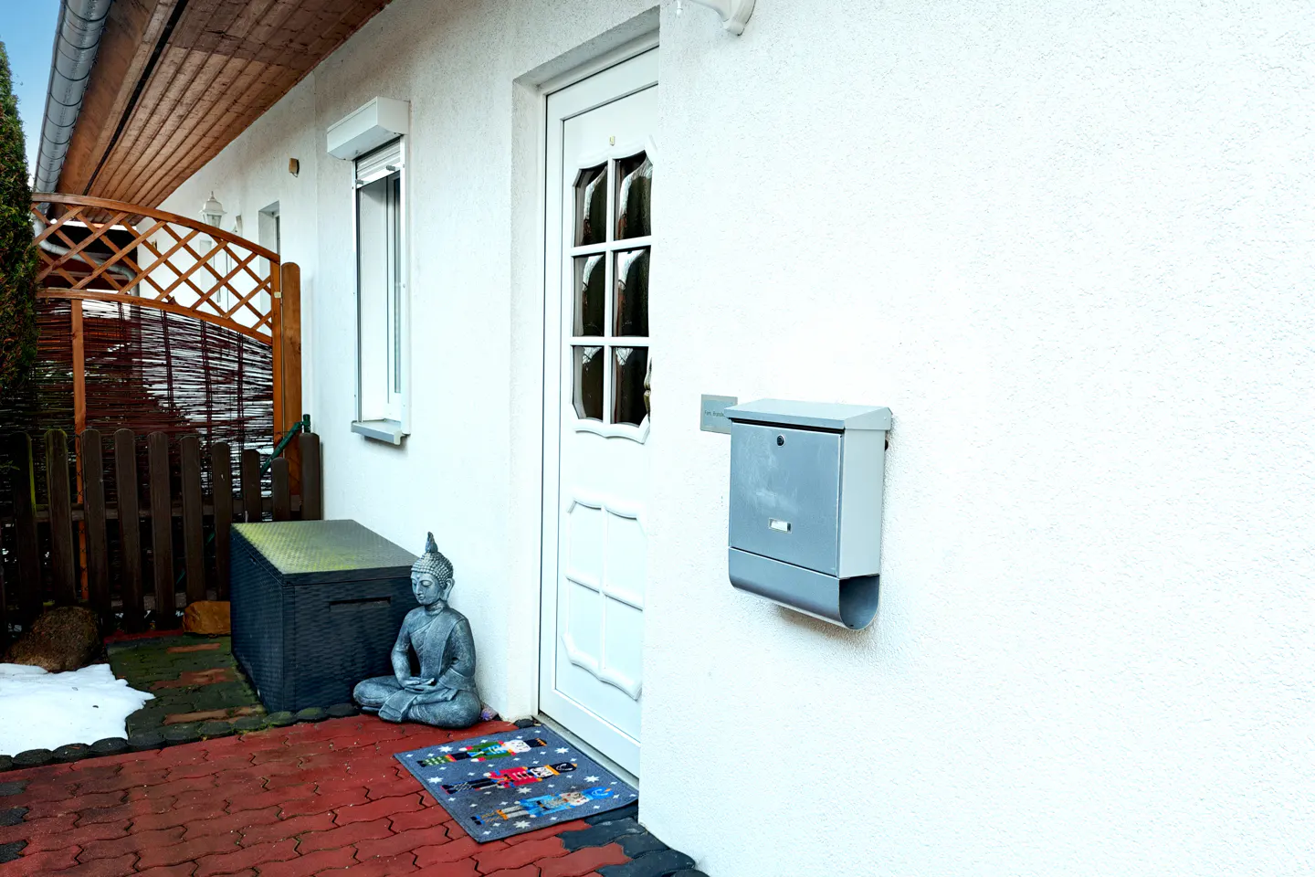Exterior view of a white house with a white door, gray mailbox, and a Buddha statue on a red brick patio.