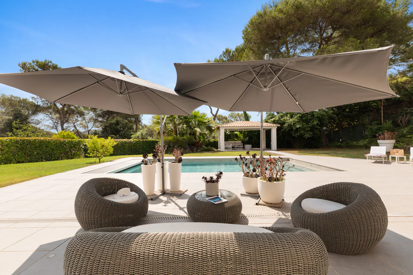 Outdoor patio with wicker furniture, gray umbrellas, and a pool. A white gazebo is in the background.