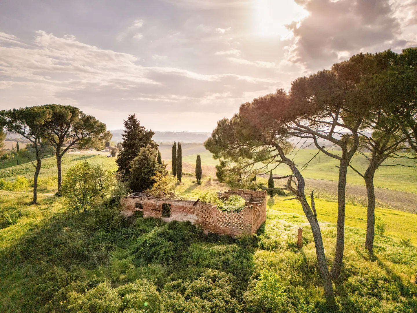 Tuscany landscape with a ruined brick building surrounded by green grass and trees under a cloudy sky.