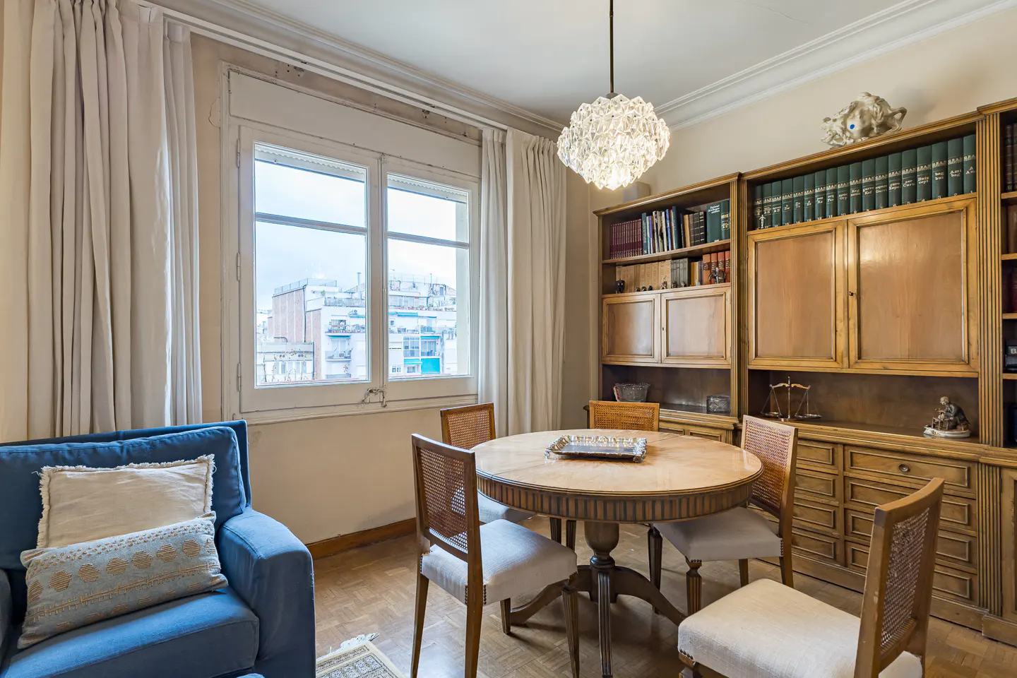 Dining area with a round wooden table, four cane-back chairs, a large wooden bookcase, and a blue armchair by the window.