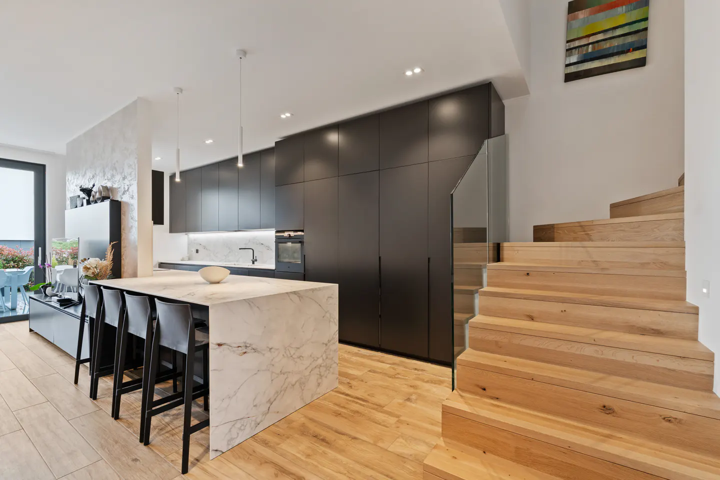 A modern kitchen with black cabinets, a marble island with four chairs, and light wood stairs.