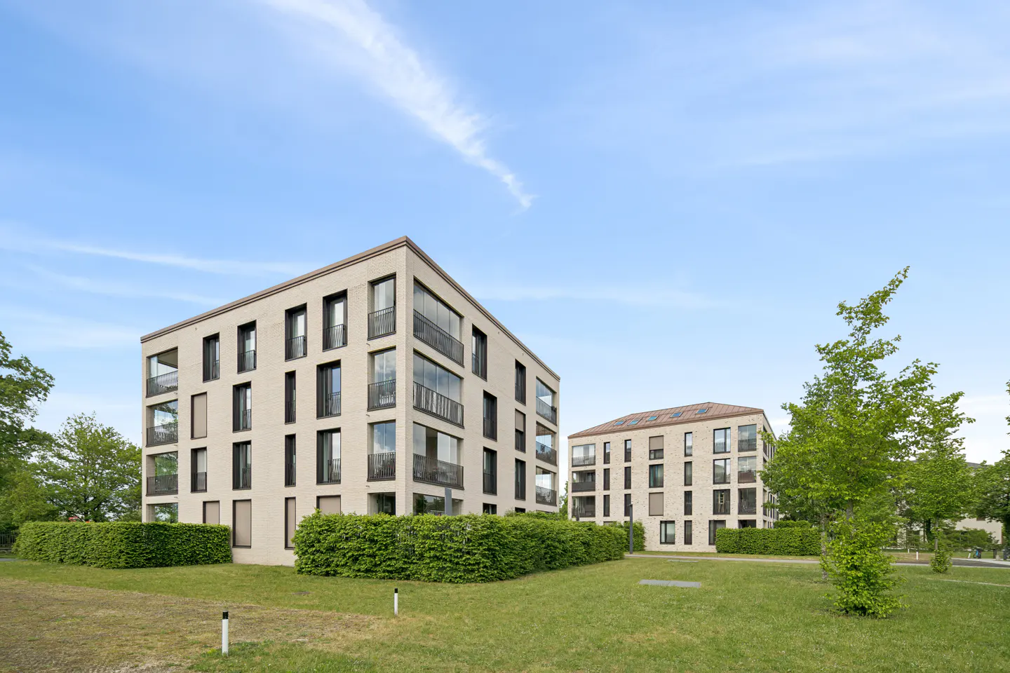 Two modern, beige apartment buildings with black-framed windows, surrounded by green lawns and hedges under a blue sky.