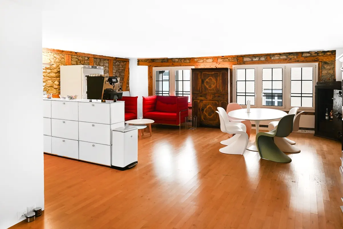 Bright, open living space with wood floors, stone walls, and exposed beams. A red sofa, white cabinets, and a round table with colorful chairs are visible.
