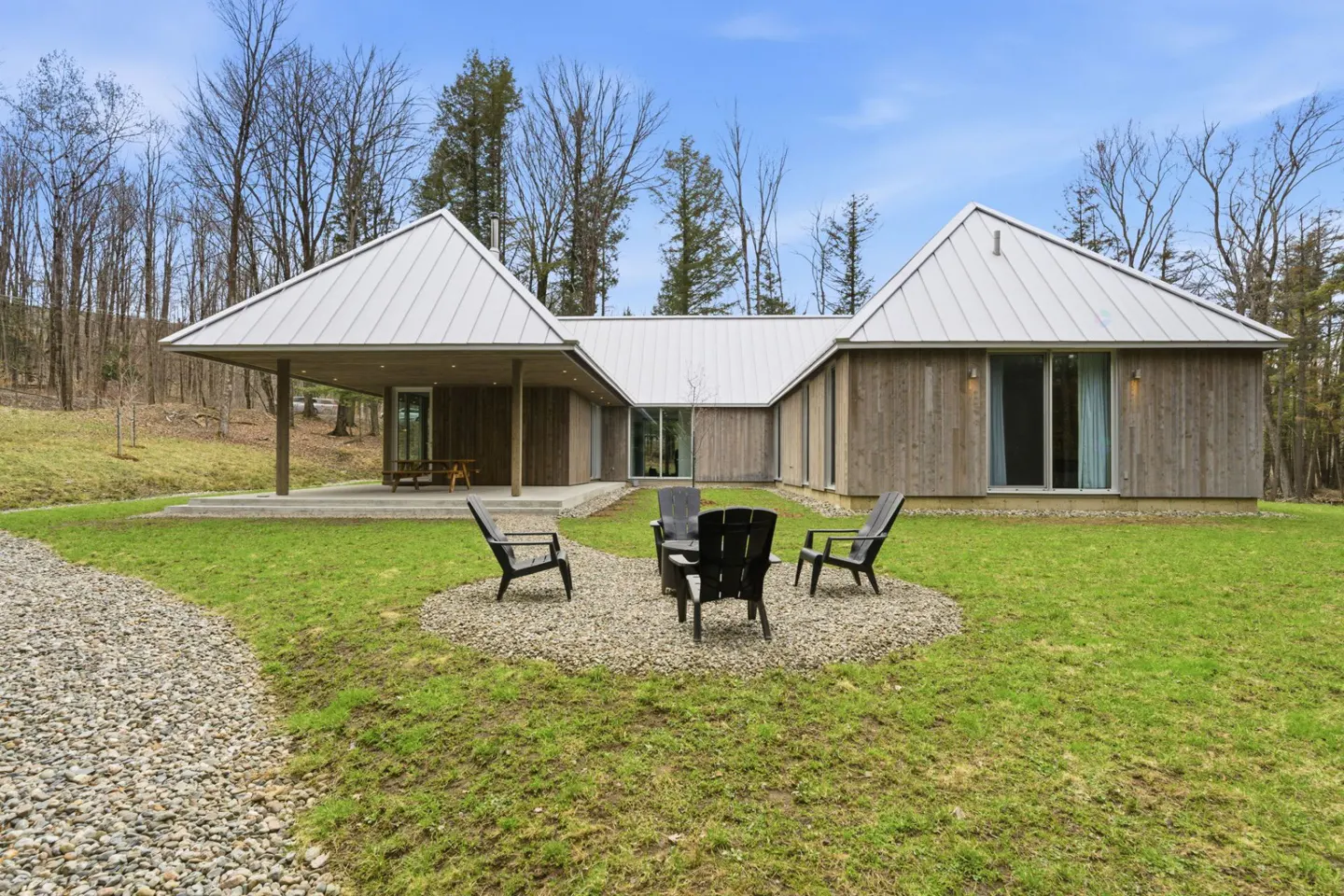 Modern wood home with a metal roof, surrounded by green grass and trees. Four black chairs sit on a gravel patio.