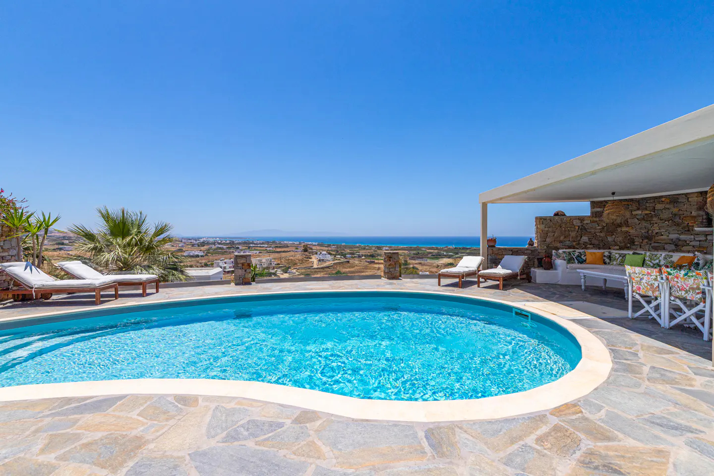 A kidney-shaped pool on a stone patio with lounge chairs, overlooking a coastal landscape under a clear blue sky.