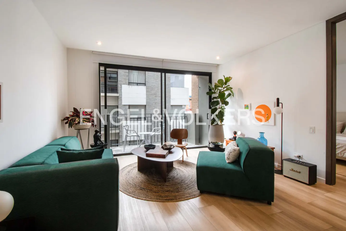Bright living room with green sofa, chair, and round coffee table on a jute rug. Balcony with chairs visible through sliding glass doors.