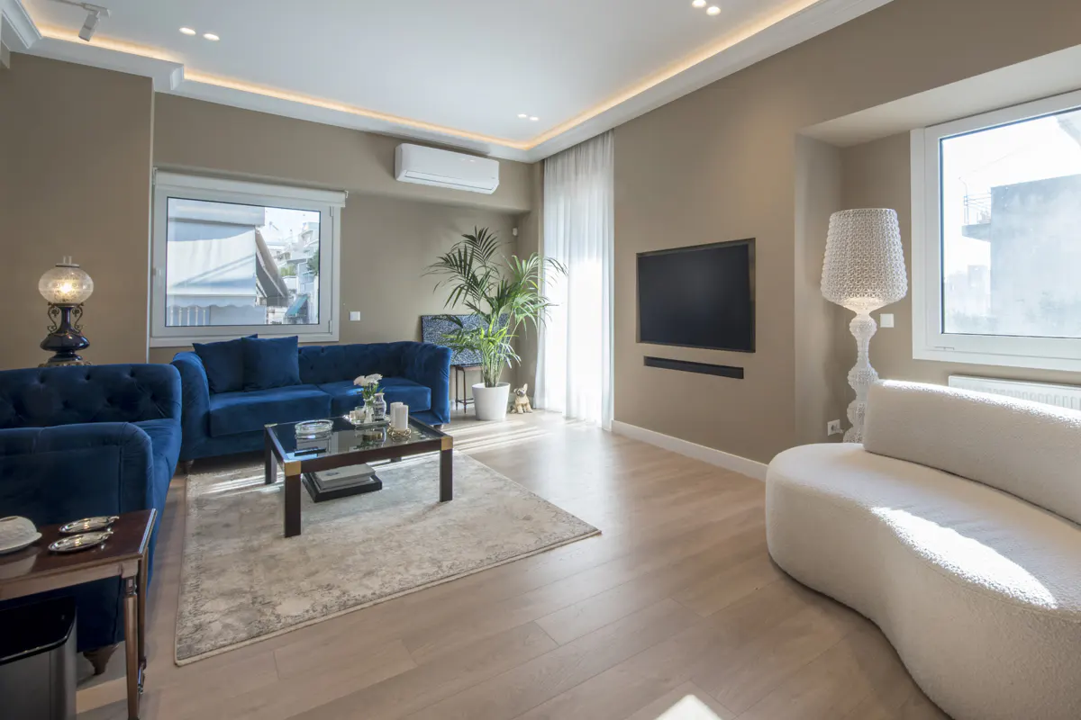 Living room with blue velvet sofas, a white curved couch, and a flat-screen TV on a beige wall. A glass coffee table sits on a patterned rug.
