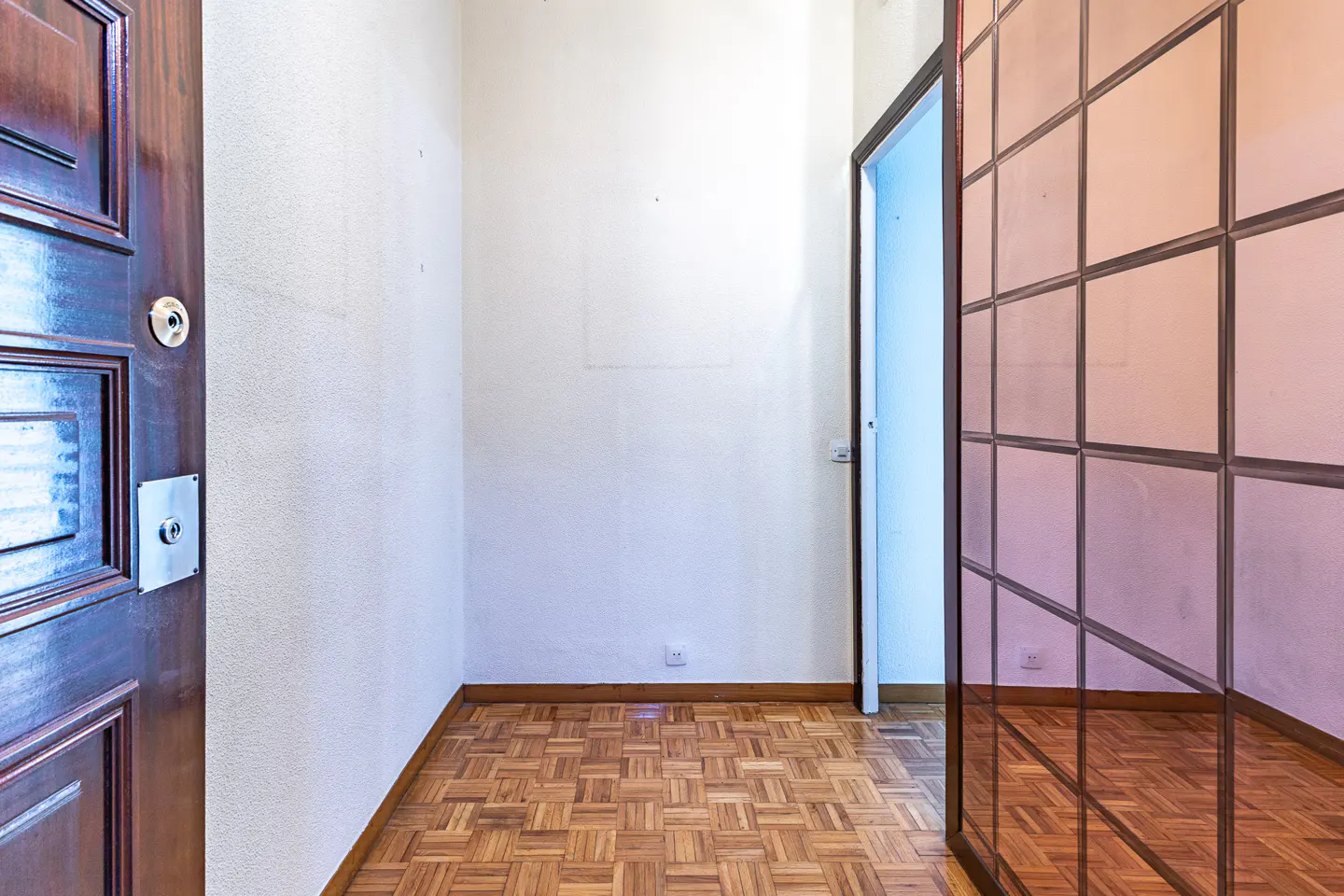 A bright room with parquet floors, white walls, and a dark wood door. A glass-paneled wall reflects the room.