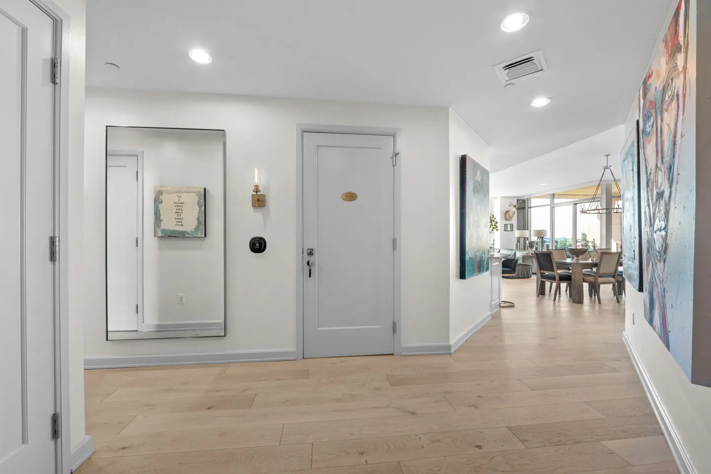 Bright hallway with light wood floors, white walls, and recessed lighting. A mirror, door, and art adorn the walls. Dining room visible in the background.