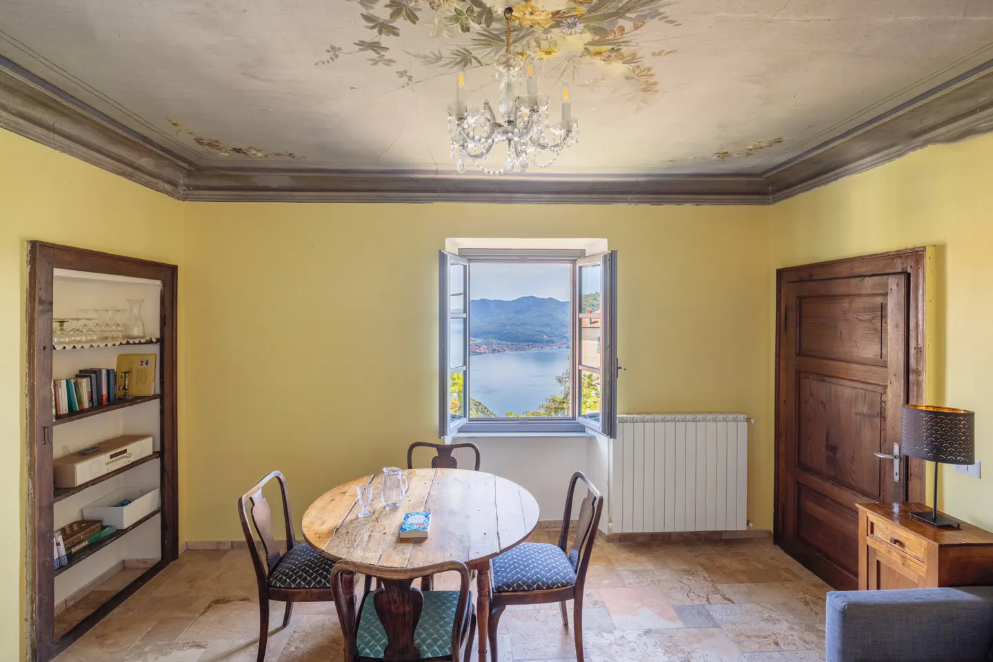 Dining room with yellow walls, a round wooden table, and chairs. A window overlooks a lake and mountains. A chandelier hangs from a floral ceiling.