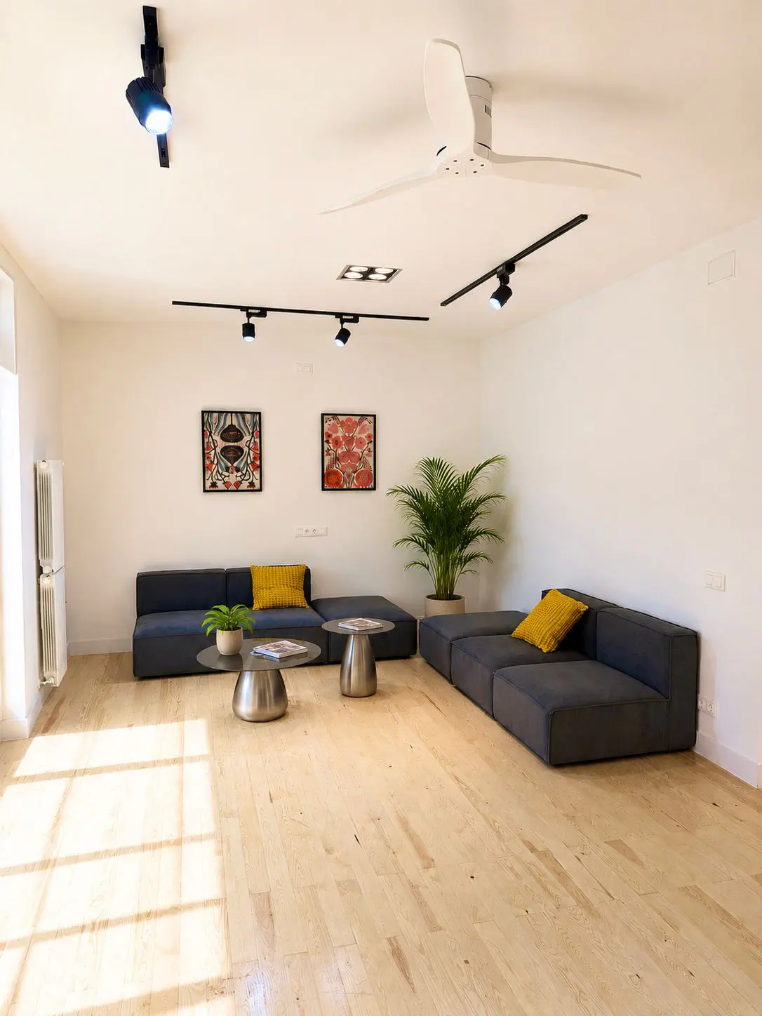 Modern living room with light wood floors, two grey modular sofas, yellow cushions, two round tables, and a large plant.