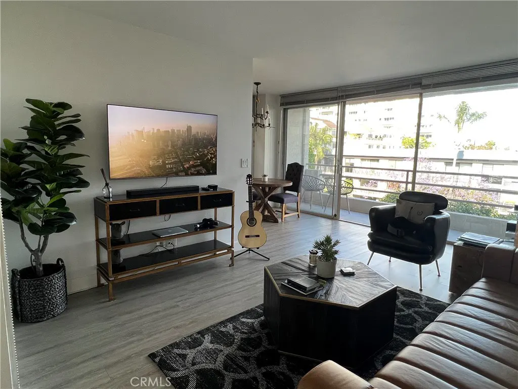 Living room with a TV, black console, guitar, table, and seating. Balcony with trees visible through sliding glass doors.