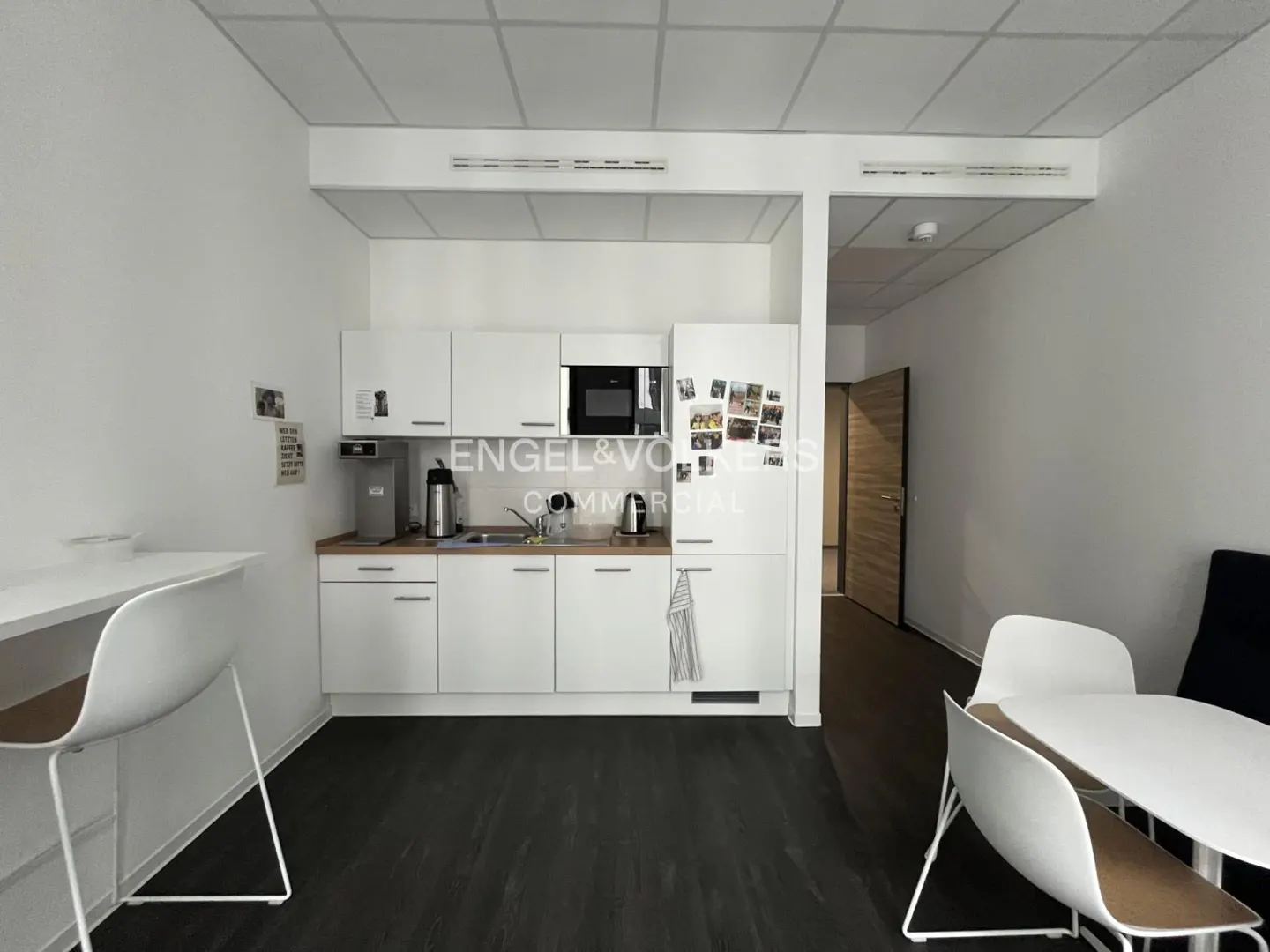 A bright office kitchen with white cabinets, a microwave, and a refrigerator. A table and chairs are in the foreground.