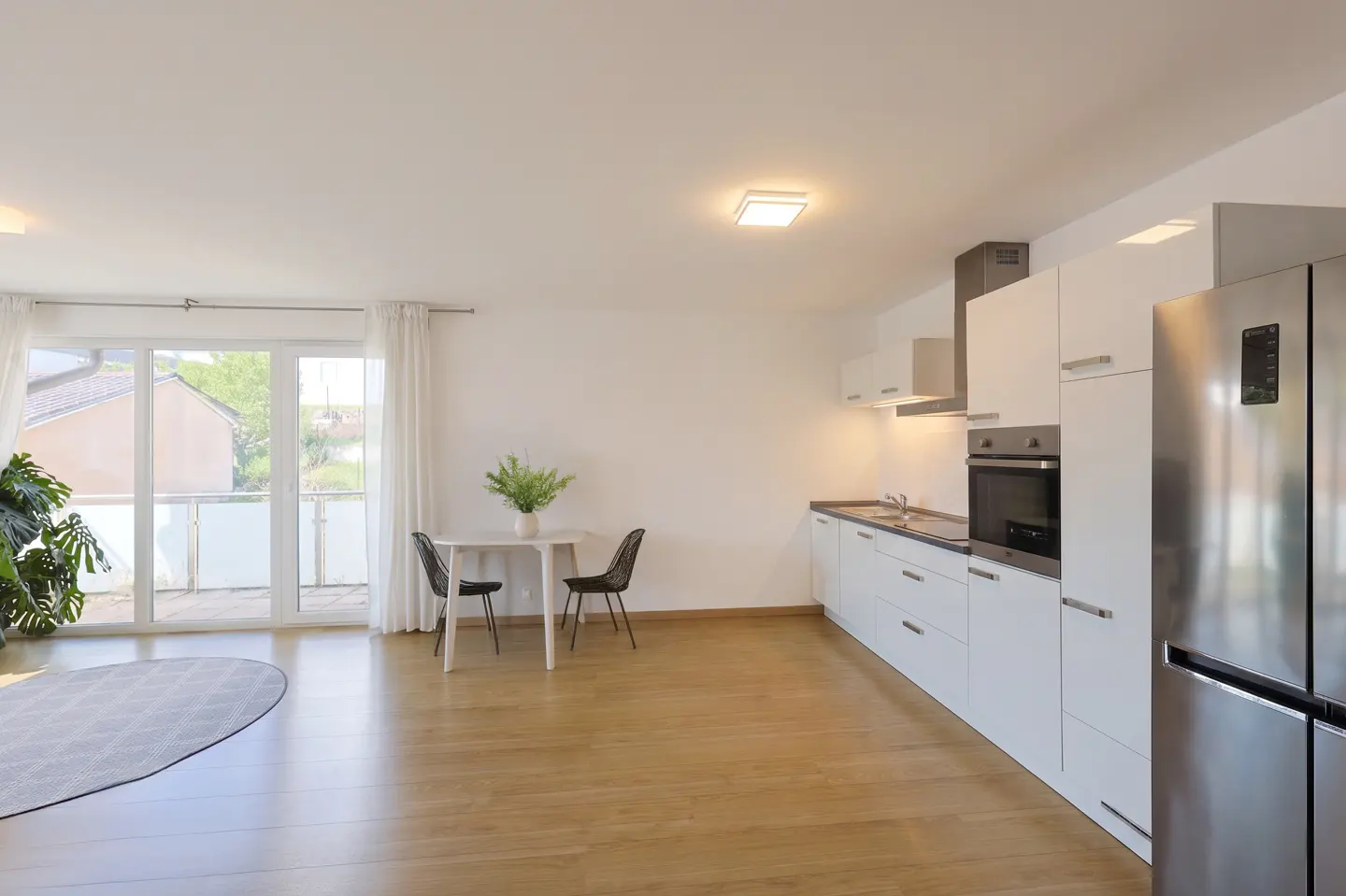 Bright, open-plan apartment with white kitchen cabinets, stainless steel fridge, and a small dining table with two chairs. Balcony doors let in natural light.
