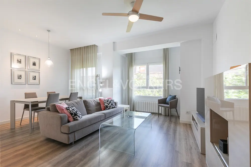 Bright living room with gray sofa, glass table, dining set, and ceiling fan. Neutral tones and natural light create a modern, airy space.