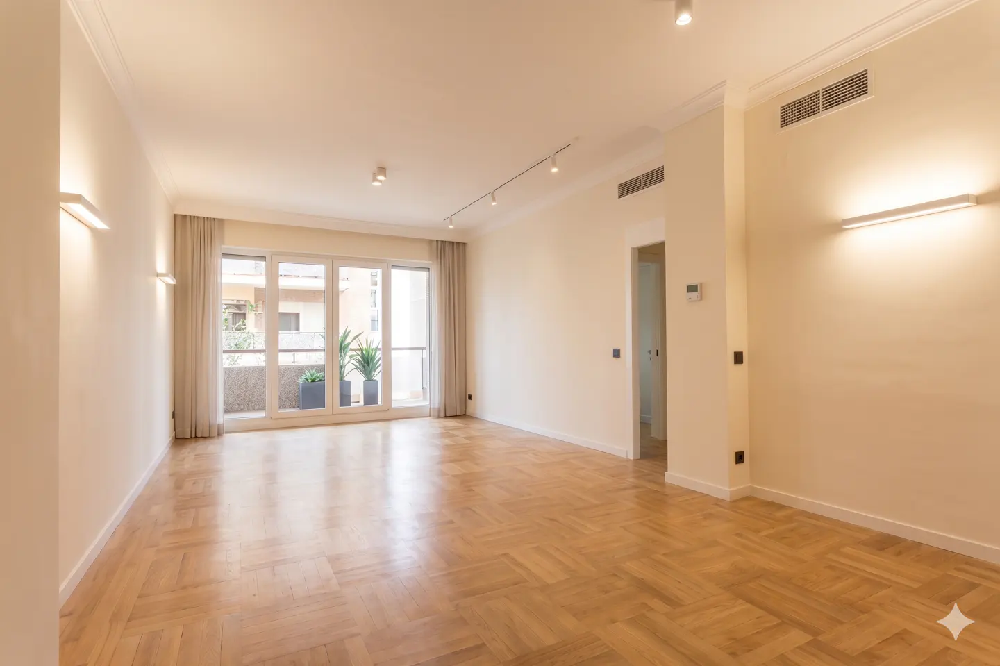 Bright, empty room with herringbone wood floors, white walls, and a balcony with plants visible through glass doors.