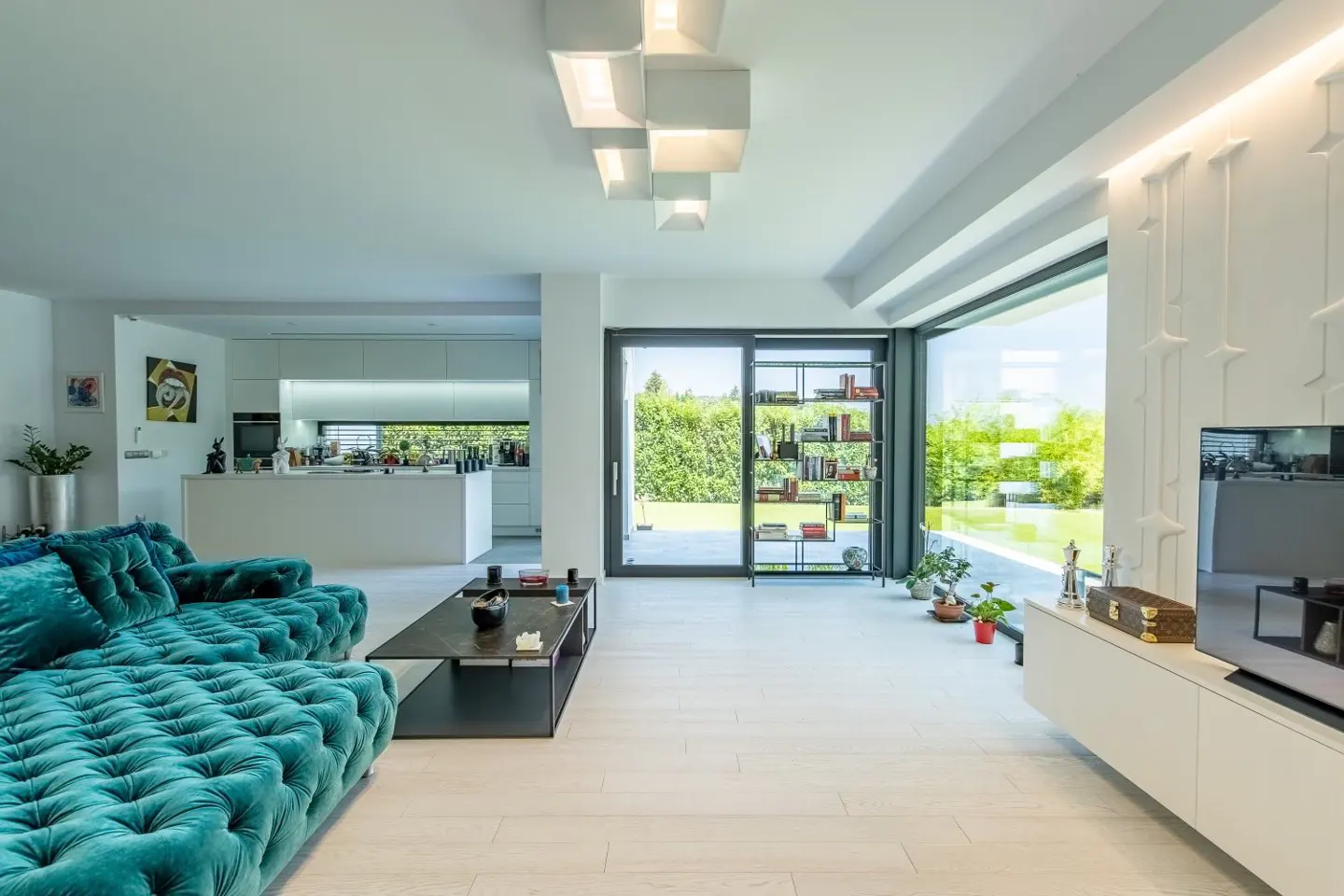 Bright, modern living room with a tufted teal sofa, black coffee table, and open kitchen. Large windows overlook a green lawn.
