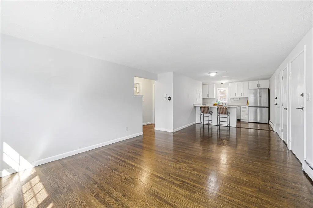 Bright, open-concept living space with hardwood floors, white walls, and a kitchen with a breakfast bar and stainless steel appliances.